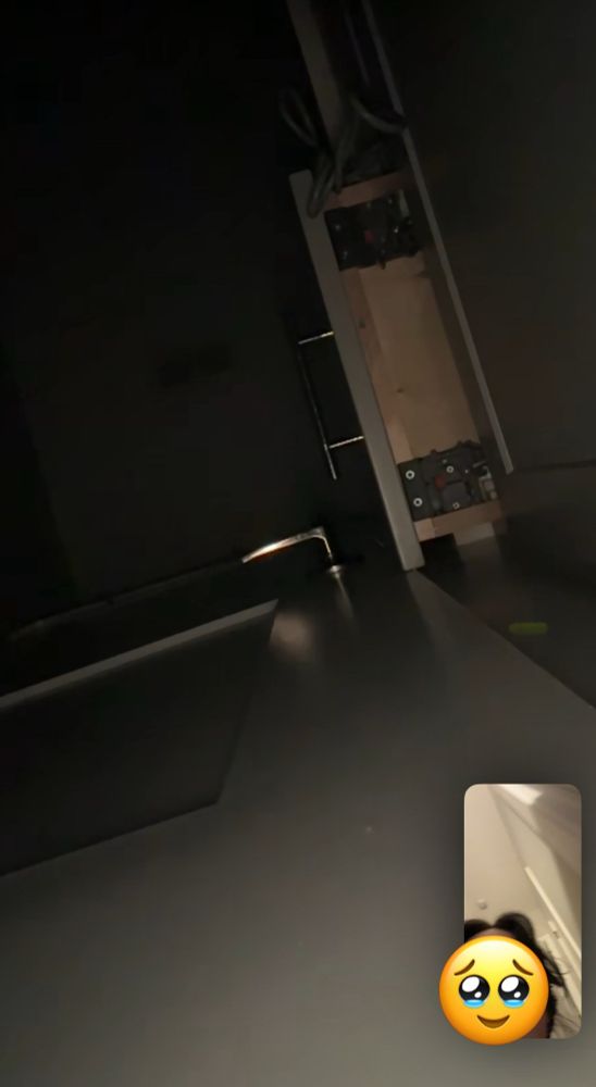 a screenshot taken from bree's phone while facetiming her roommate's phone. the roommate's phone camera is facing up illuminating a dark bathroom, showing the underside of a slightly open drawer that is preventing the door from being opened. in bottom corner, bree (face covered with a teary eyed smiley emoji) can be seen watching her roommate's progress in closing the drawer. 
