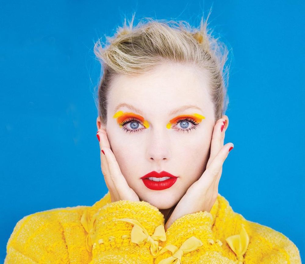 Taylor Swift dressed in yellow, with her hair pulled back, her hands on either side of her face with red fingernails. 
her lips are painted red, her eyeshadow is orange and yellow.