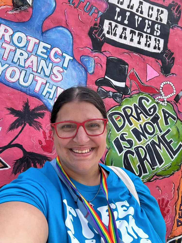 A white woman with red glasses and a rainbow lanyard standing in front of a colorful mural that says Protect Trans Youth, Black Lives Matter and Draf is not a Crime.
