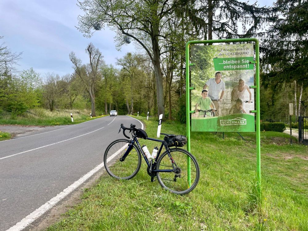 Rennrad lehnt am Landkreisschild der Uckermark neben der Straße, dahinter Bäume