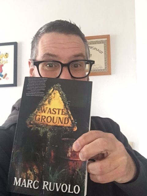 A dumb guy with glasses holding up his dumb cosmic horror book WASTE GROUND. He wishes more people would read it, but, you know...
