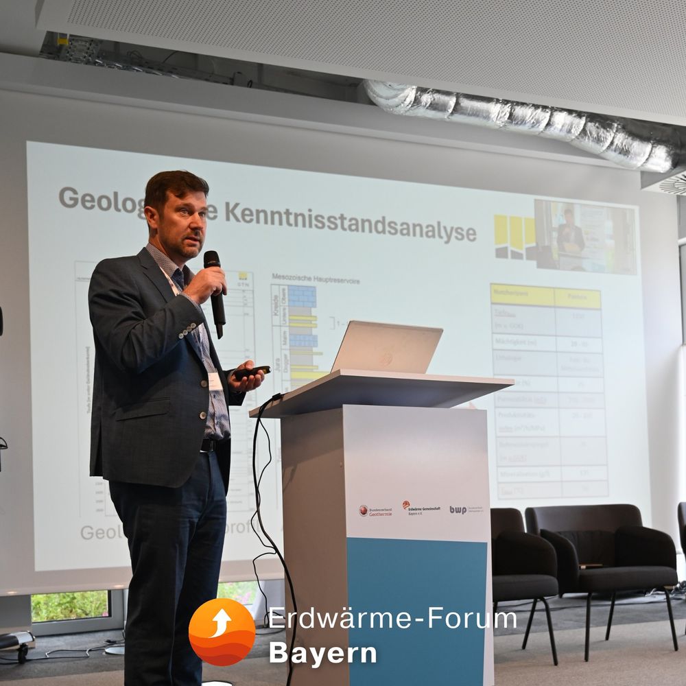 Ein Mann in Anzug und Hemd steht auf einer Konferenzbühne und hält mit Mikrofon in der rechten Hand einen Vortrag. Vor ihm steht ein Rednerpult mit Laptop, im Hintergrund ist eine Präsentationsfolie mit dem Titel „Geologische Kenntnisstandsanalyse“ zu sehen. Die Folie enthält geologische Grafiken und eine Tabelle. Auf dem Pult sind Logos der Veranstalter sichtbar, darunter das des Bundesverbands Wärmepumpe (BWP) und der Geothermie-Gemeinschaft Bayern. Unten links im Bild befindet sich das Logo des Erdwärme-Forum Bayern mit stilisiertem Aufwärtspfeil in Orange.