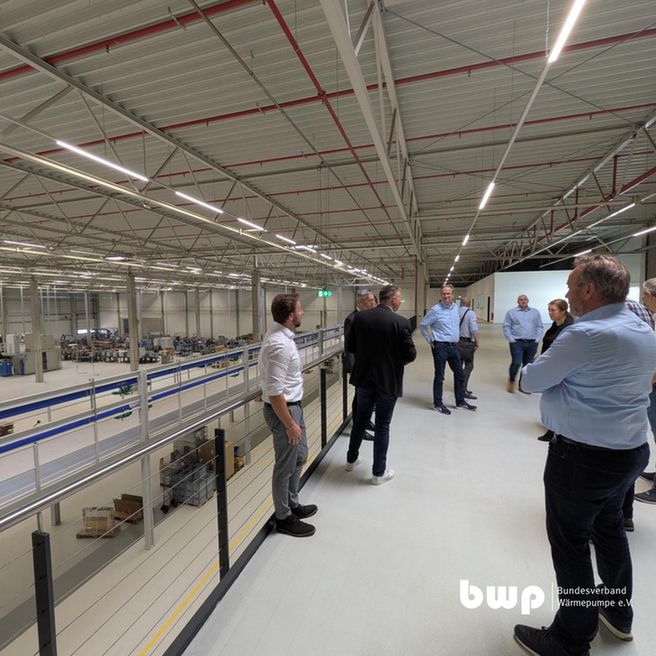 Teilnehmer einer Delegation stehen auf einer Galerieebene mit Blick in eine große, moderne Produktionshalle. Die Halle ist hoch, sauber und hell, mit offenen Tragwerkskonstruktionen und industriellem Bodenbelag. Eine Gruppe von Personen – einige in Hemd oder Sakko – unterhält sich und blickt teilweise in Richtung Kamera.  Das BWP-Logo ist unten rechts eingeblendet.