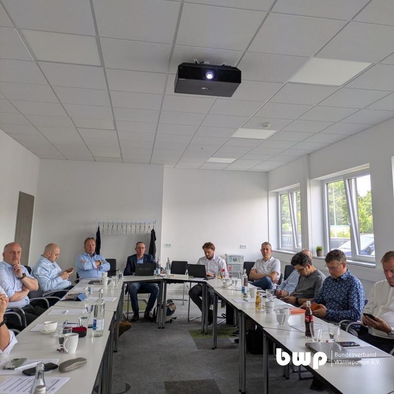 Mehrere Personen – überwiegend Männer in Hemd oder Sakko – sitzen um einen rechteckigen Konferenztisch in einem hellen Besprechungsraum. Einige arbeiten an Laptops, andere schauen auf Mobilgeräte oder hören zu. Im Raum stehen Wasserkaraffen, Kaffeetassen und Unterlagen. An der Decke ist ein Beamer eingeschaltet. Im Hintergrund erkennt man große Fenster mit Tageslicht. Unten rechts ist das Logo des Bundesverbands Wärmepumpe (bwp) eingeblendet.
