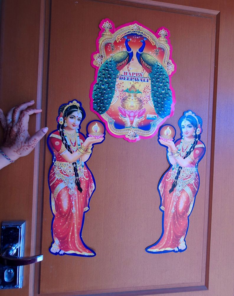 A henna-ed hand rests against a brown front door with three decorative signs on it. At the top is an elaborate one with decorative Indian elements and Tamil text (தீபாவளி வாழ்த்துக்கள்) on it, over two large peacocks. Below them is the text Happy Deepavali, over floral confetti and a traditional Tamil prayer item of a gold vessel with leaves and a coconut, on a lotus held by jewelled hands.

Below that and to either side are identical mirrored signs. Each one shows a woman with long braided hair, dressed in a traditional sari and lots of jewellery, holding a lotus on which sits an oil lamp with a large flame. 