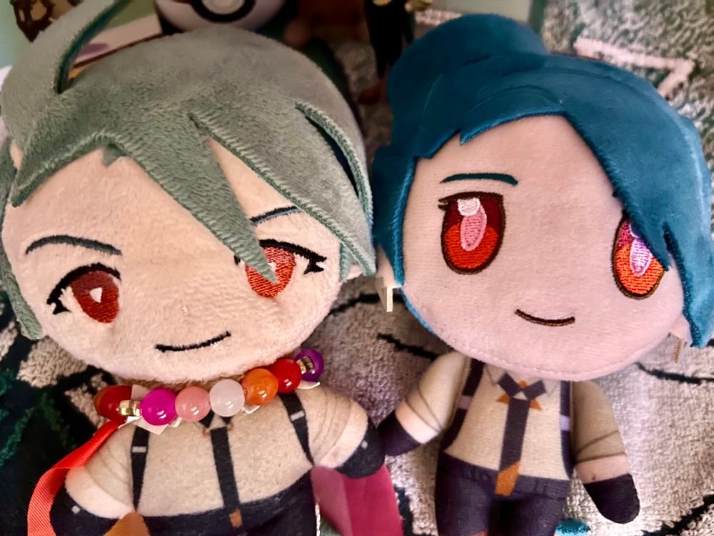 Two official Rika plushies, side by side. The left one has a lesbian pride necklace and flag as a cape