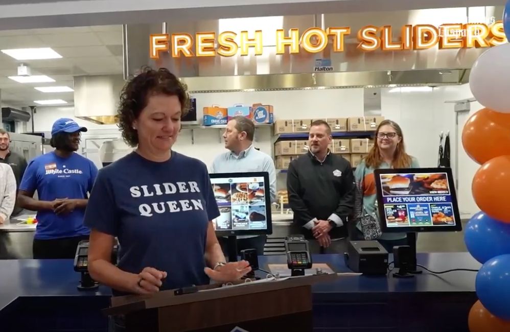 A woman wearing a t-shirt that reads "Slider Queen," standing behind a podium in a White Castle restaurant, no doubt about to say something momentous. There is a lighted sign behind her which reads "Fresh Hot Sliders." There are red, white, and blue balloons to the right. 