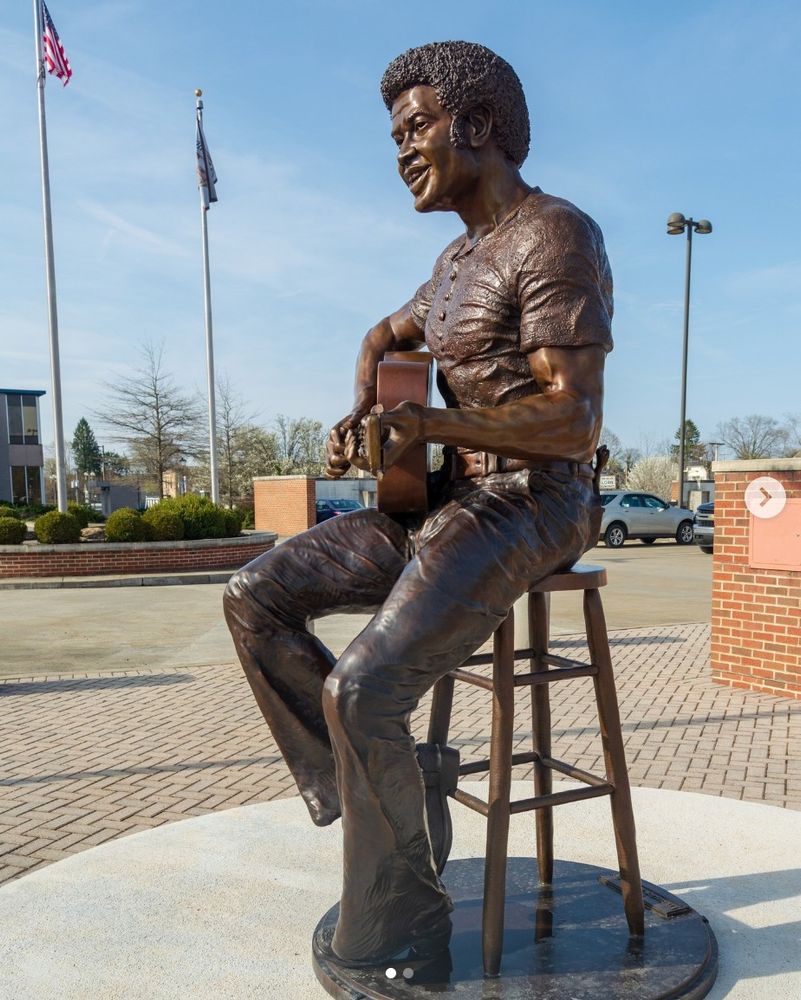 A bronze statue of Bill Withers, sitting on a stool, playing guitar in an outdoor public plaza