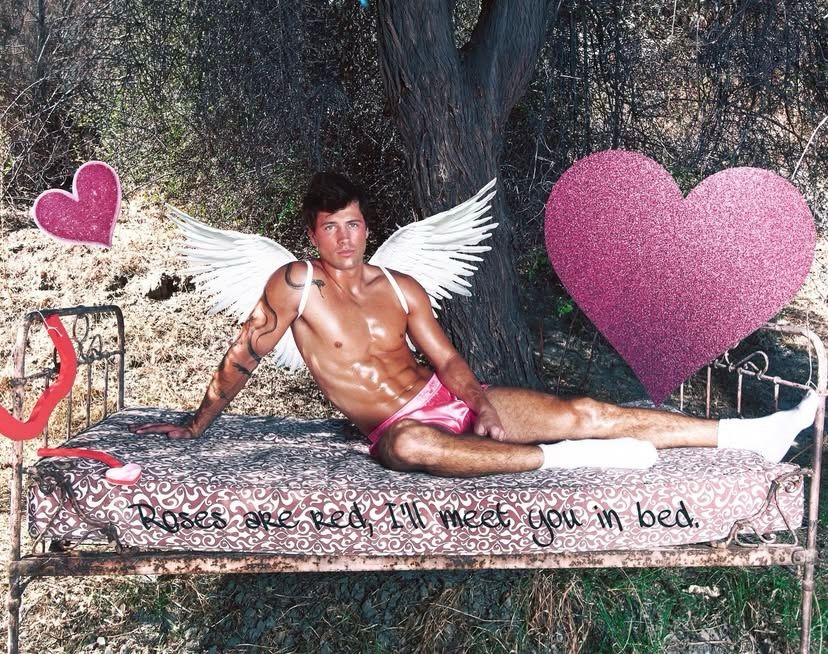 rob shirtless with angel wings. the text on the bed says “roses are red, i’ll meet you in bed.”