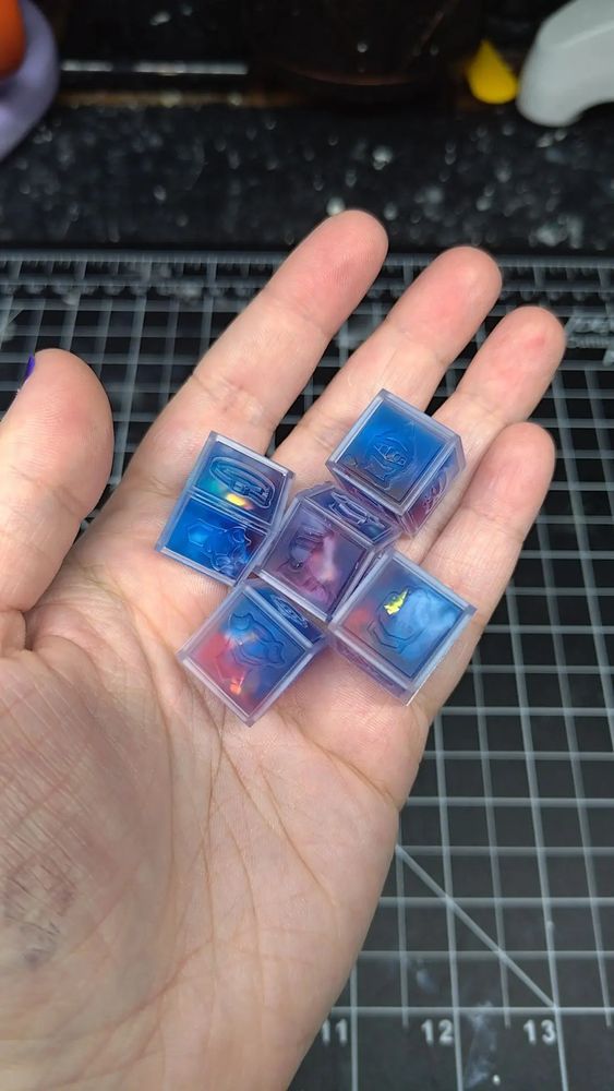 5 d6 cubed dice that uses iconography for hit locations; helmet/head, belt/waist, glove/hand, foot, and chest. The resin used is clear, blue, and red, with white to imitate clouds, and mylar in the center.