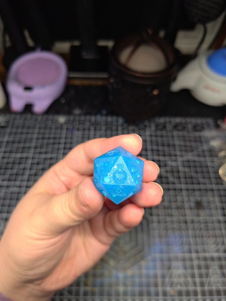 A death save d20, with the icon of a potion bottle and heart facing the camera. The overall d20 is very blue, with reflective material in the d20.