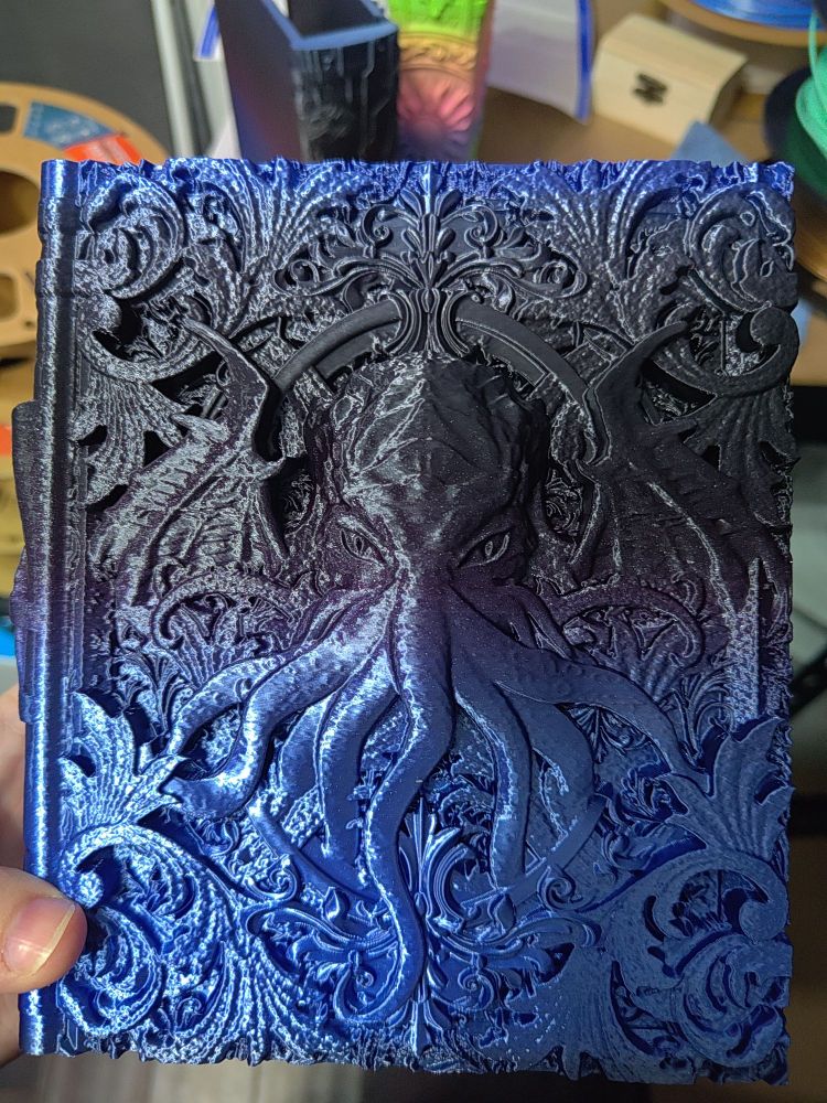 A 3D printed storage tome for not only TTRPG players, but you can use them for anything! The outside tome has a unique design, while the faux pages hide the secret storage compartments. This tome has a 3D image of a Chathulu's head (an alien face with tentacles where his nose and mouth should be), with demon wings and barque textures on the cover. The filament used is a gradient filament that goes from black to blue to purple, and has sparkly glitter mixed into the filament.