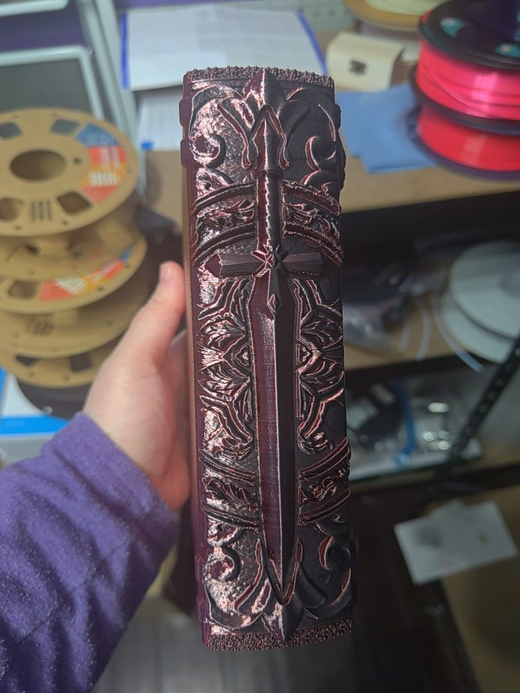 A 3D printed tome with a sword on the front and additional barque textures surrounded the sword. This is the spine of the tome and is mostly covered by the sword.