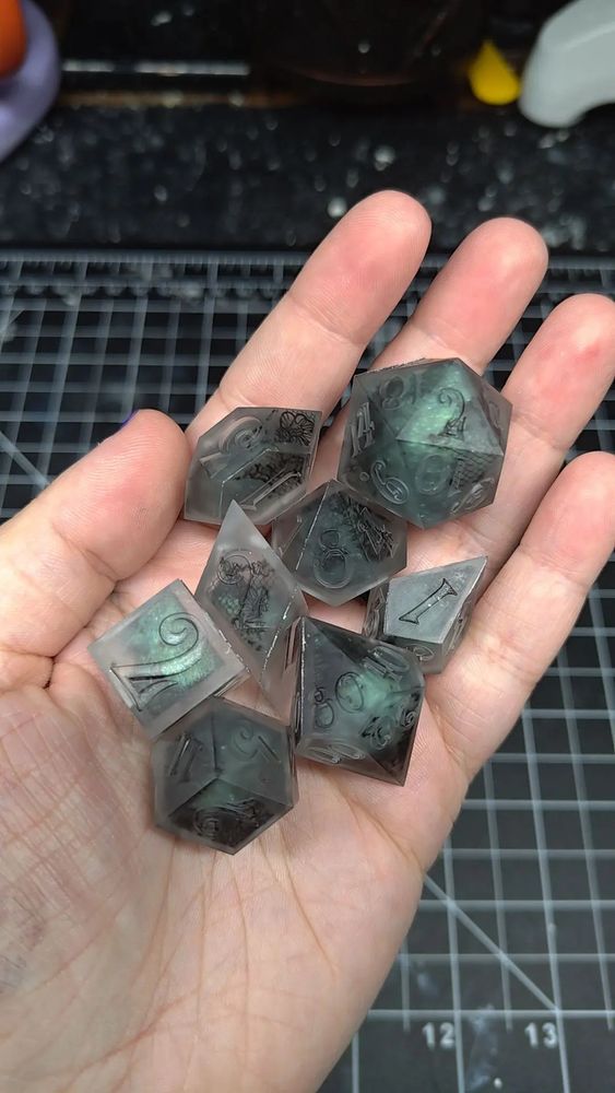 8 Polyhedral dice of varying shapes and sizes. the font used is a whimsical curly font. the resin used is clear, transparent black and ghost green.