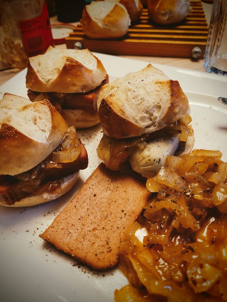 Auf einem Teller liegen 3 Laugenbrötchen. Zwei davon sind mit veganen Fleischkäse und eins mit Weißwurst gefüllt. Zusätzlich ist auf jedem Brötchen eine Portion Schmorzwiebeln und daneben liegt ein weiteres Stück Fleischkäse und Zwiebeln.