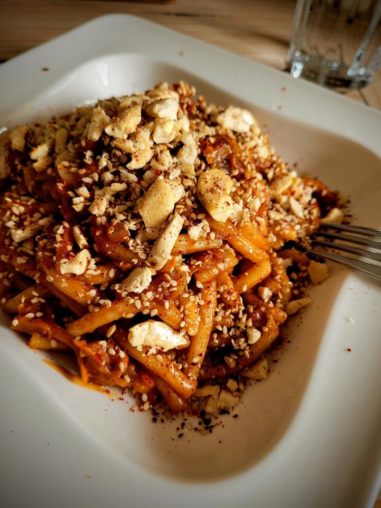 Ein weißer Teller mit Udon in braun-oranger Erdnuss-Chili-Sauce. Als Topping sind gerösteter Cashewbruch und Sesam oben drauf.