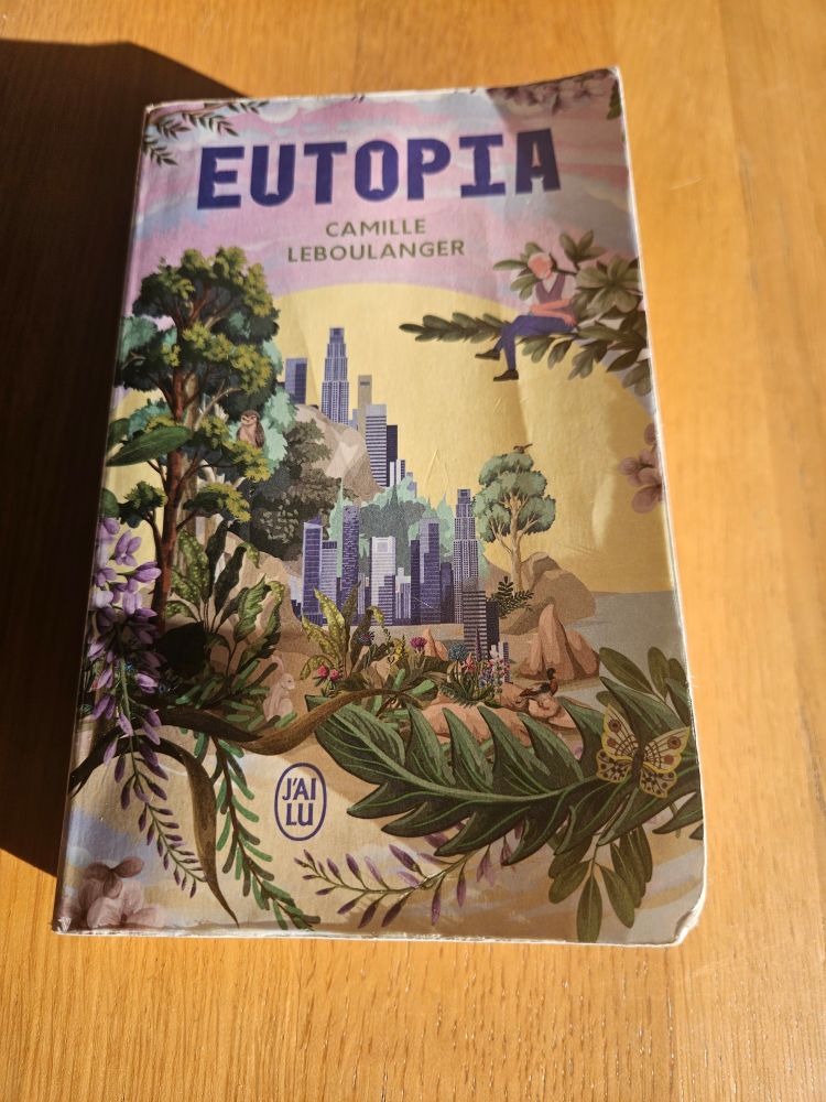 La couverture du livre "eutopia" de camille leboulanget aux éditions j'ai lu. On y voit des gratte-ciel au bord de l'eau avec des arbres et des plantes au premier plan et un demi soleil en arrière plan. Sur une branche en haut à droite, un personnage âgé.  