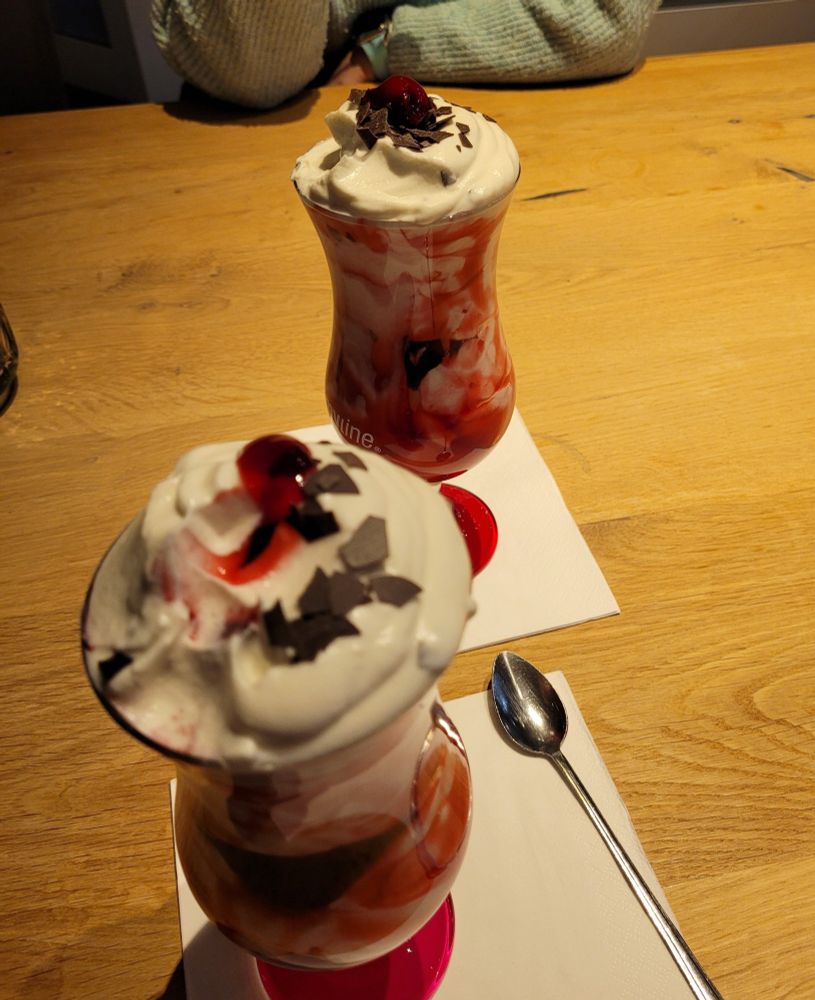 Zwei Stilgläser auf einem Tisch. In den Gläsern befindet sich ein weißer Shake mit rot von Kirschen durchzogen. Darauf Schokoflocken und eine Kirsche