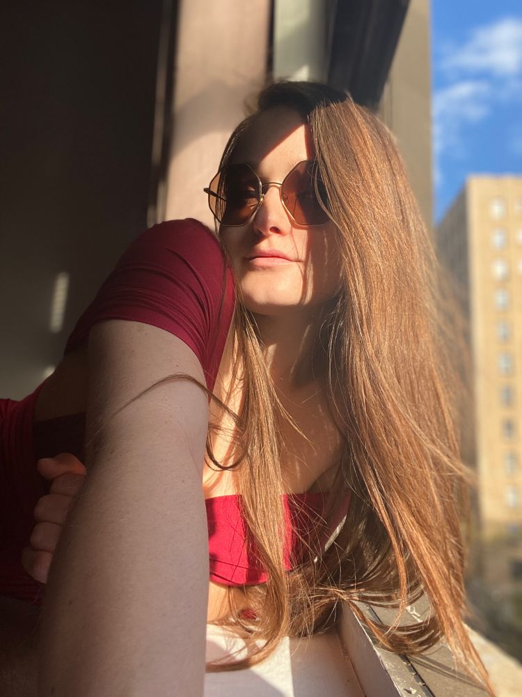 A white woman in her late twenties takes a selfie of her leaning on a windowsill. The window is pushed all the way open and her long auburn-ish hair catches the sunlight and trails out over the threshold. It’s the longest her hair has ever been. She’s wearing a red wine colored bridesmaid dress and oversized octagonal sunglasses. The sky beyond is blue and speckled with fluffy clouds.
