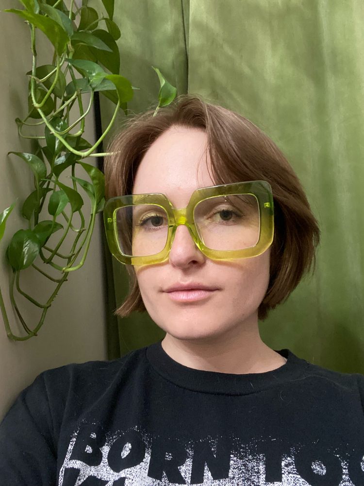 Photo of me - a 30 year old white woman. My hair is medium brown and hits just at my chin. It’s freshly cut and very cute. My glasses are oversized lime green square frames and I’m wearing a black shirt with text visible that says “born too.” I’m standing in front of a green curtain and a white dresser with a tendril of a pothos plant trailing just behind my shoulder.