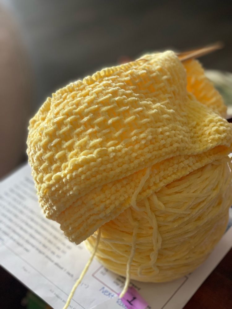 A shot of a work-in-progress knitting project. There is the beginning of a light yellow blanket perched on top an attached cake of yarn. There are about four inches of fabric made, which has a basket-weave-like texture.