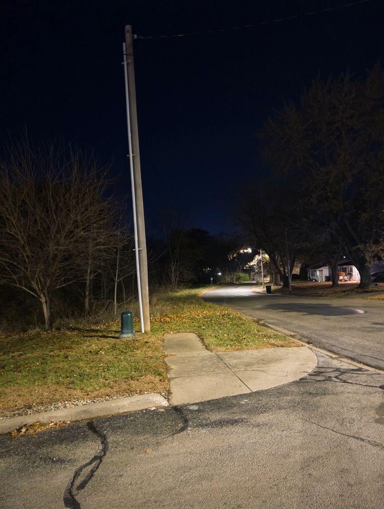 A photograph taken from the top of a hill at night, facing down said hill. A road goes down the hill. It’s lit with street lights. There’s a forest to the left, some trees and houses to the right. A dark blue sky is visible. There’s a subtle soft glow in the distance.