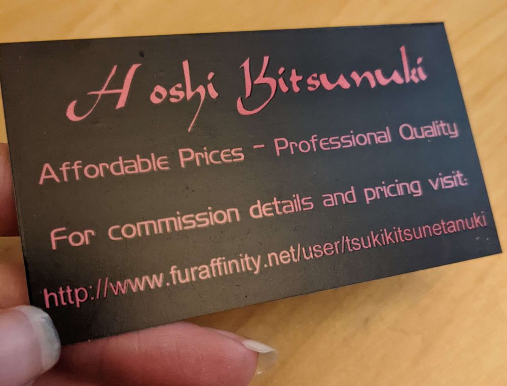 A photo of an old business card for our commission business which reads "Hoshi Kitsunuki / Affordable Prices - Professional Quality / For commission details and pricing visit: http://www.furaffinity.net/user/tsukikitsunetanuki

Card is reflective black with pink lettering.