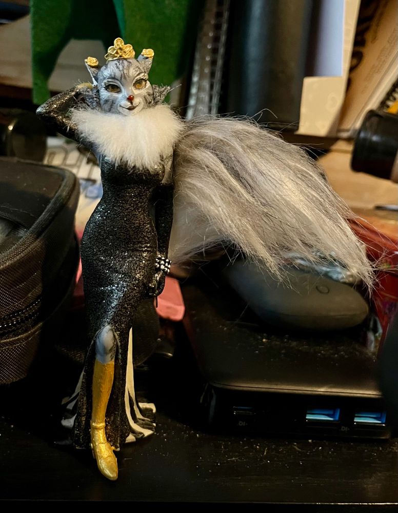 An anthropomorphic cat figure that has been hand painted and modified to look like my original character Sycavity. She has a golden calf and foot as well as a golden hand partially obscured by this photograph. She has a long faux fur tail hand painted at the top, neatly trimmed white faux fur at the neck, and painted faux fur coming off her cheeks. She is wearing a black shimmery evening gown.
