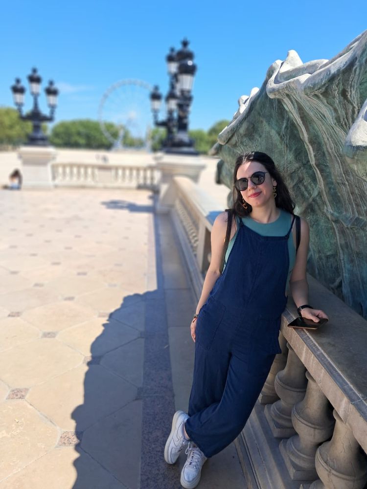 Foto de mí misma con un peto vaquero suelto, top celeste, botines blancos y llevo gafas de sol. De fondo se ve una noria. 