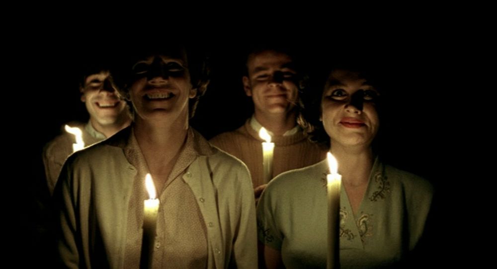 four faces wearing white jackets or sweaters lit from below by candles