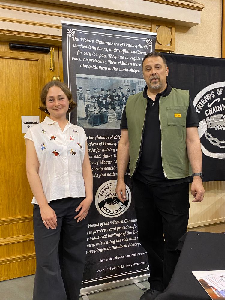 Polly and Stuart stand in front of a banner dedicated the the women chainmakers and their battle for decent pay