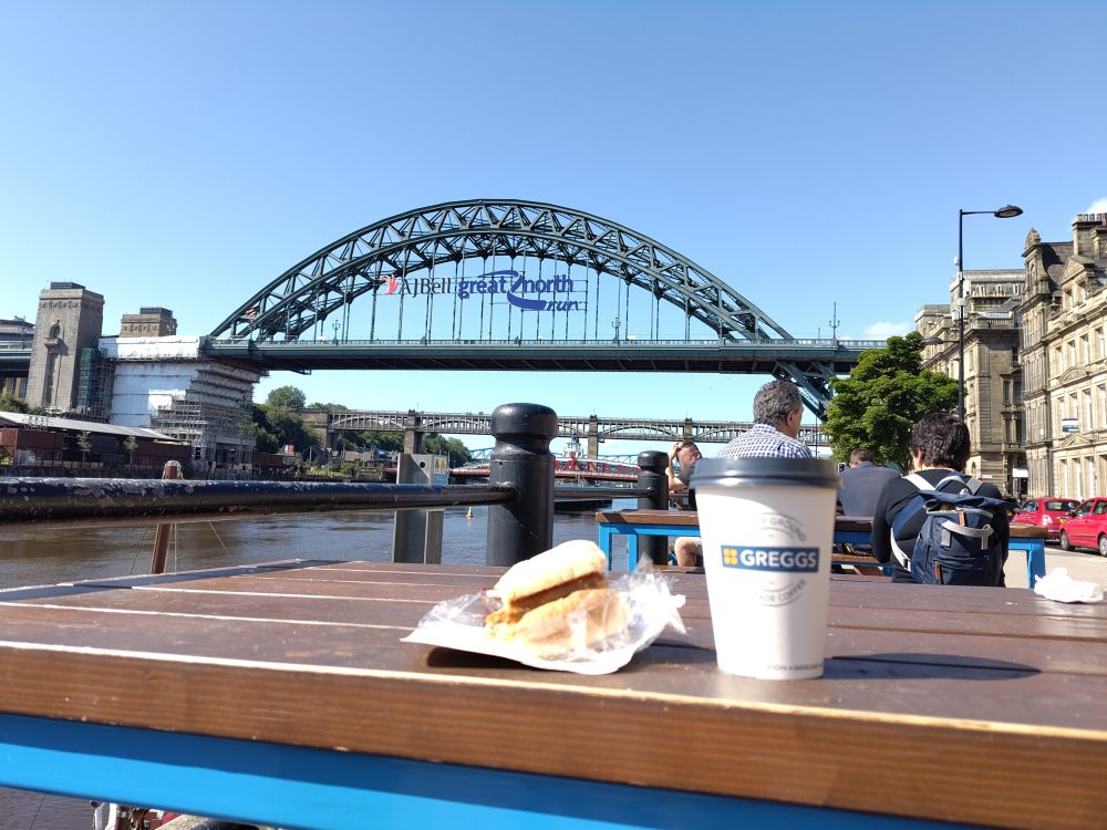 A Greggs coffee and breakfast roll on table with Tyne Bridge in the background