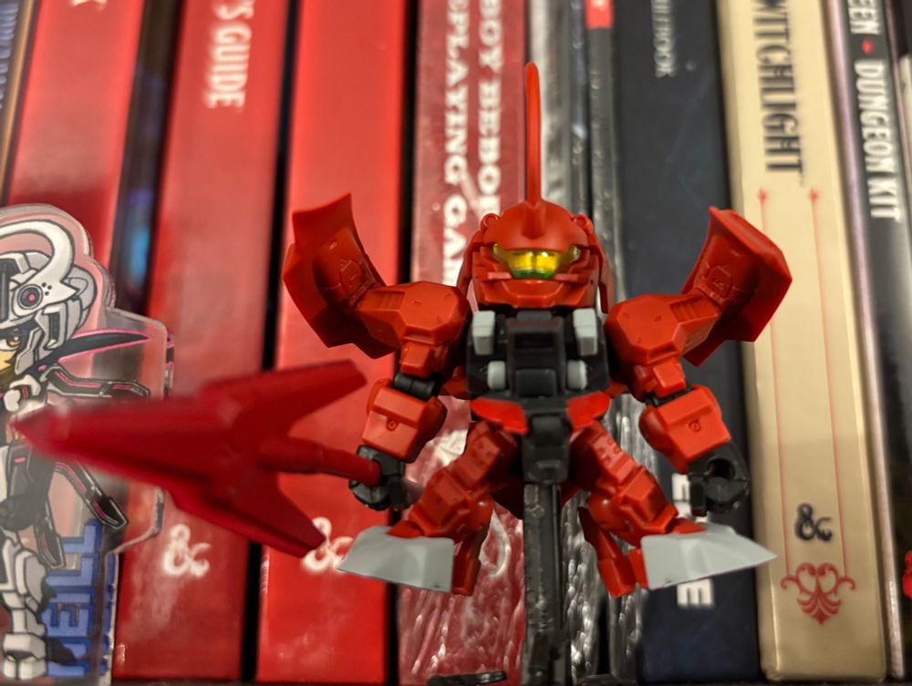 Photo of a darilblade gunpla gashapon kit with ttrpg books behind, up close at an angle
