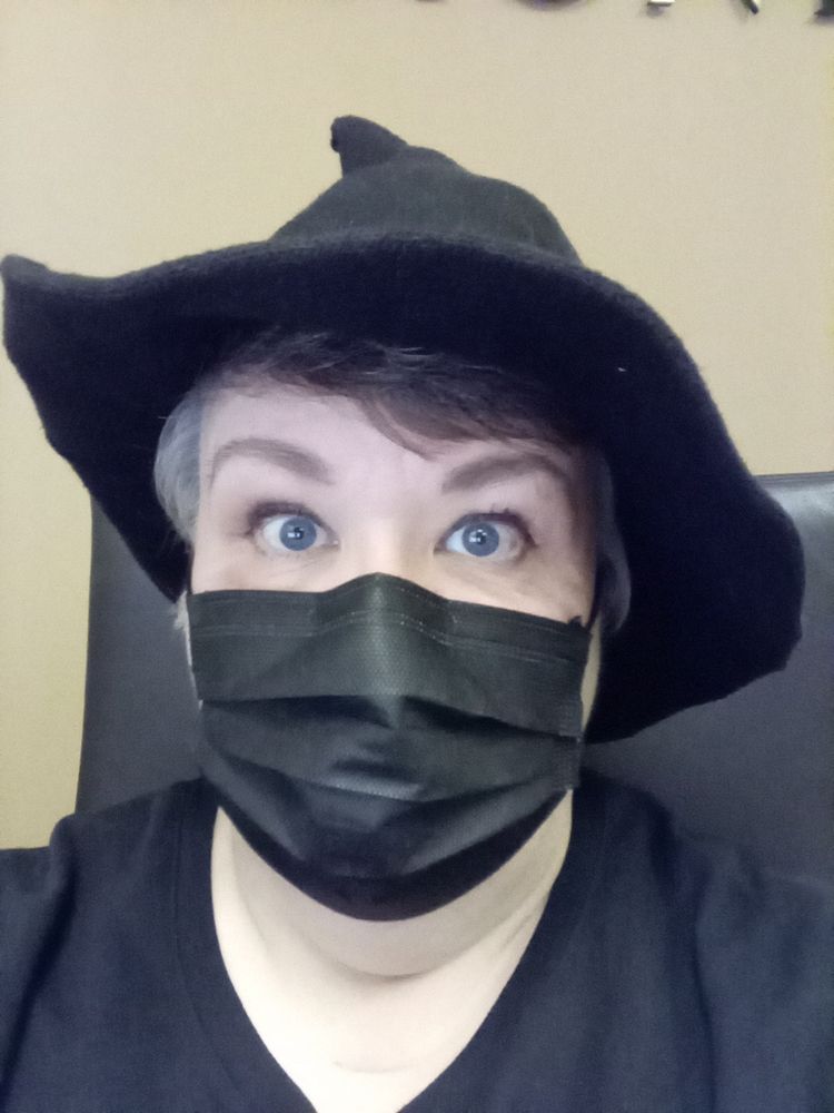 A middle-aged white woman wears a black surgical mask and a black witch hat.