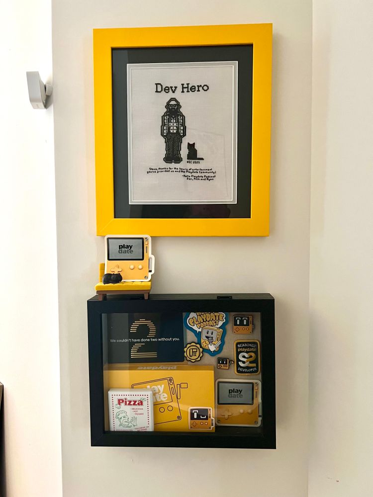 A shadow box mounted on a wall containing a lot of bits and bobs related to the Playdate. Above it hangs a cross stitch of me from Post Hero. 