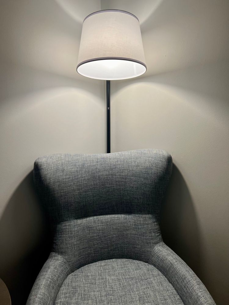 A chair in the corner of a room with a floor lamp behind it, angled directly over the chair. 