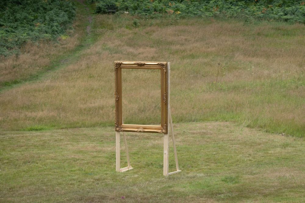 A large gilt picture frame is mounted upright on wooden supports and standsby itself on rough grass. There is nothing in the frame.