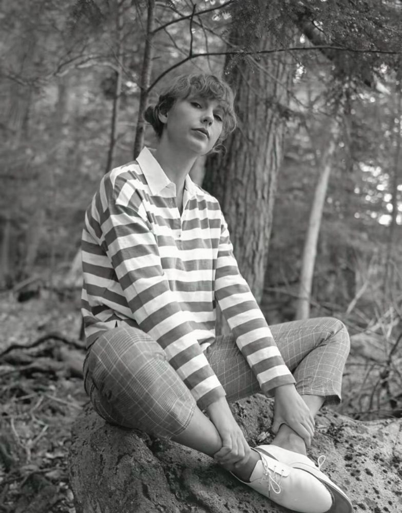 a picture of Taylor Swift sitting in the woods in a striped shirt and plaid pants from folklore era looking gay af 