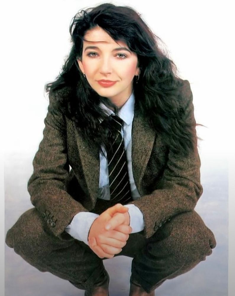 A photograph of Kate Bush with her hair down wearing a light brown wool suit, shirt, and striped tie 