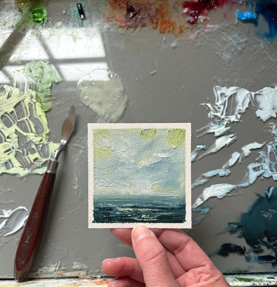 Photo of a hand holding an original tiny seascape oil painting by Tisha Mark, "Tiny Dream No. 2502" 3"x3" oil on Arches oil paper (2025). Tiny textured seascape painting with a blue, green, and white sky over a deep blue ocean. Colors and light from the sky reflect in the water below. A paint palette with puddles of oil paint and a palette knife are visible in the background.