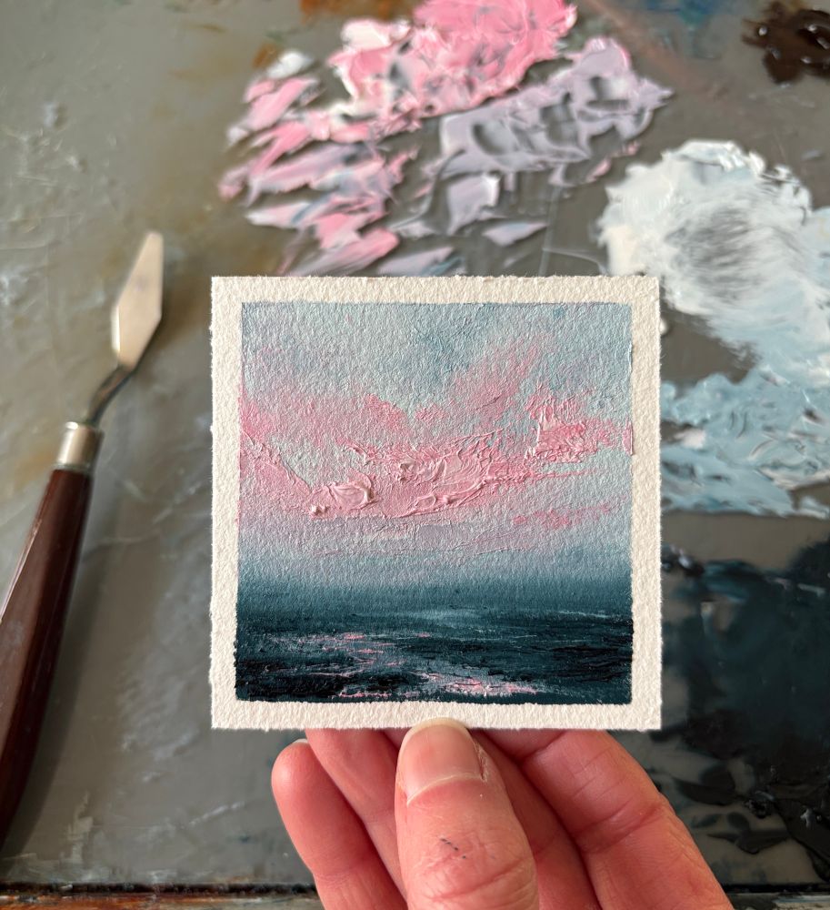 Photo of a hand holding an original tiny seascape oil painting by Tisha Mark, "Tiny Wild Sunset No. 2507" oil on Arches oil paper, 3"x3" (2025). Seascape painting with pink cloud formations in a light blue sky over a dark blue sea with a rocky shore. Light from the sky reflects in the water below. A paint palette with puddles of paint and a palette knife are visible in the background.