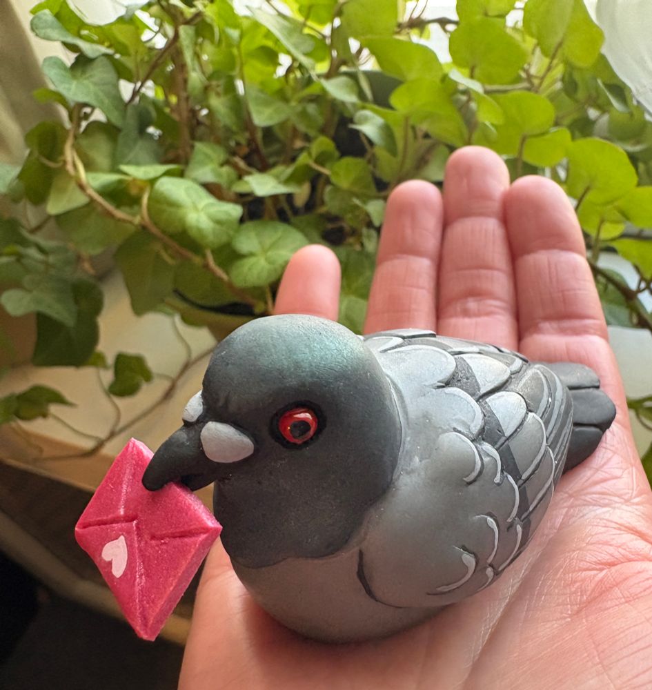 Photo of an adorable resin pigeon sculpture with a valentine in their beak! Pigeon sits in the palm of my hand, a green plant is in the background. 