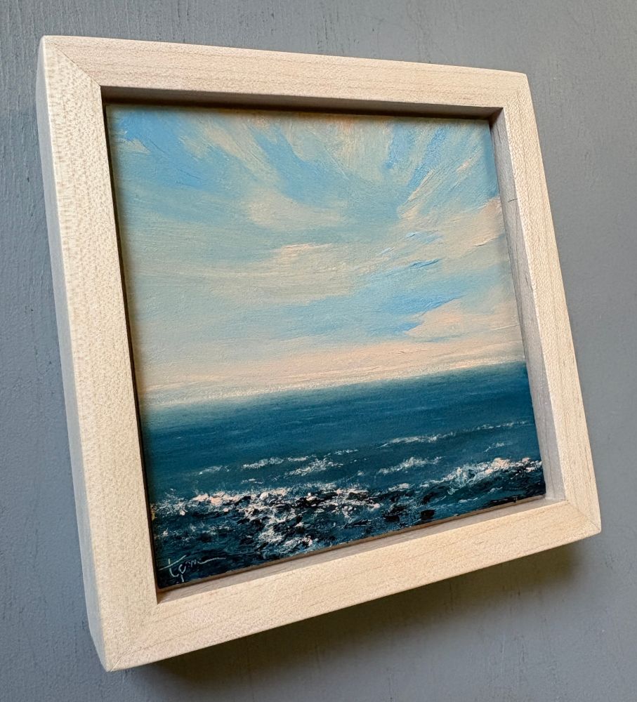 Original seascape oil painting by Tisha Mark, "A Little Hopeful Light No. 2501" 4"x4" oil on Ampersand Gessobord (2025), shown in a maple frame at an angle from left to show paint textures and depth of frame. Seascape painting with the hopeful light of an orange-toned sunrise or sunset sky over a deep blue sea.