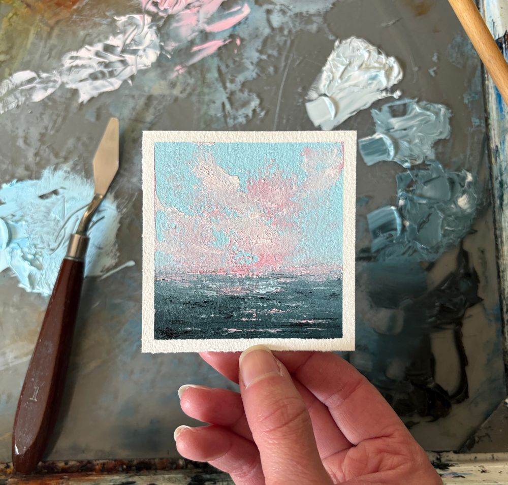 Photo of a hand holding an original seascape oil painting by Tisha Mark, "Tiny Summer Sunrise No. 2505" oil on Arches oil paper, 3"x3" (2025). Seascape painting with a blue sky with pink sunlit cloud formations over a dark blue sea with a rocky shore. A paint palette with puddles of oil paint and a palette knife are visible in the background.