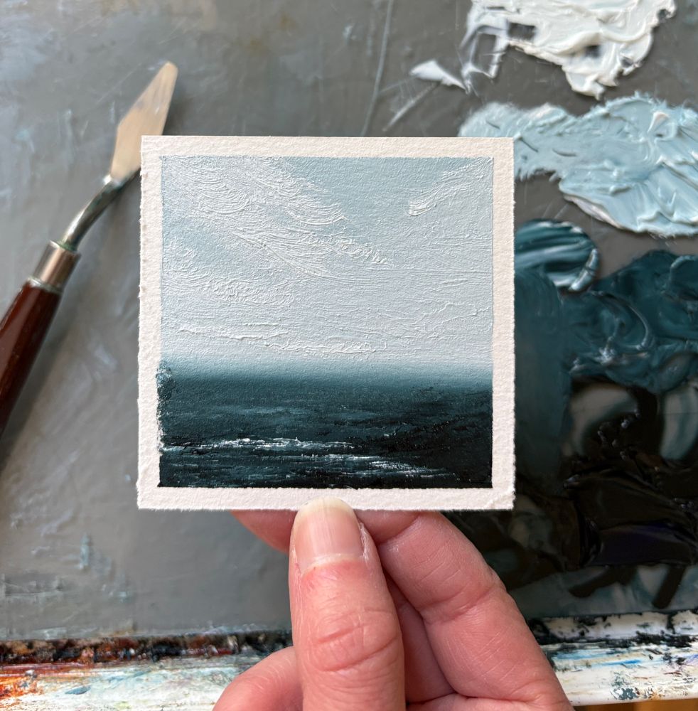 Photo of a hand holding an original tiny seascape oil painting by Tisha Mark, "Soothing Sea No. 2502" 3"x3" oil on Arches oil paper (2025). The painting is a tiny textured seascape with gentle white atmospheric clouds over a deep blue sea. A paint palette with puddles of oil paint and a palette knife are visible in the background.