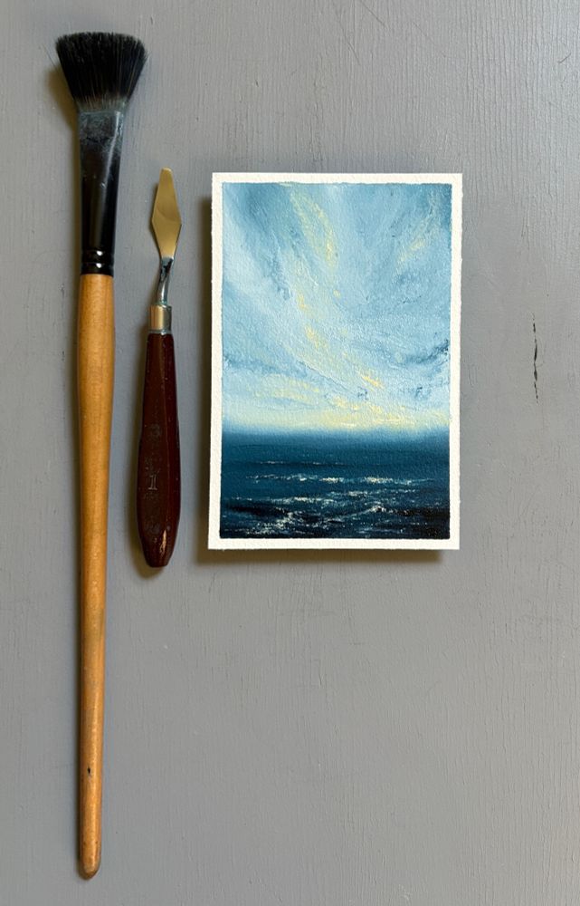 Original seascape oil painting by Tisha Mark, "Hopeful Morning No. 2502" 6"x4" oil on Arches oil paper (2025), shown with a paintbrush and palette knife for scale. Textured seascape painting with white atmospheric clouds touched by yellow sunlight in a blue sky over a deep blue sea. Yellow sunlight from the sky reflects in the water below.