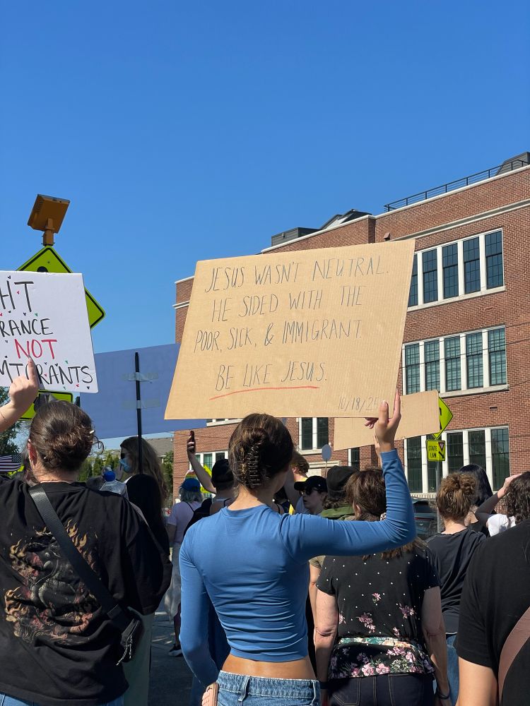 Single protestor in blue shirt & jeans holding cardboard box sign that reads “Jesus Wasn’t Neutral. He Sided With The Poor, Sick, & Immigrant. Be Like Jesus.”