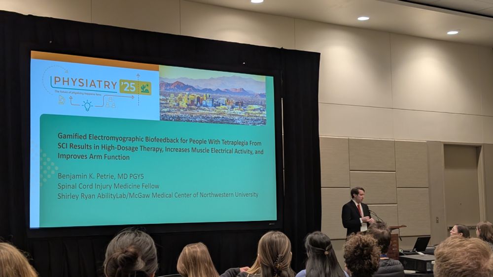 Photo of Ben Petrie standing in front of his slide title "Gamified Electromyographic Biofeedback for People with Tetraplegia