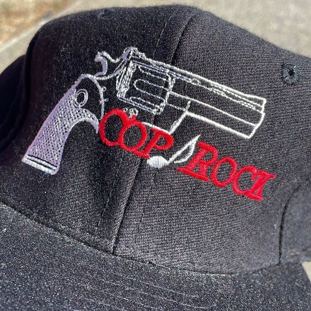 Black baseball cap featuring a stitching of a gun with a music note and text that reads COP ROCK