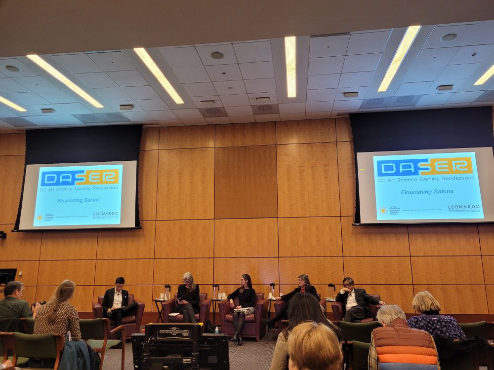 A panel of scientists in a meeting room with wooden paneling and two screens with slide presentations being projected.